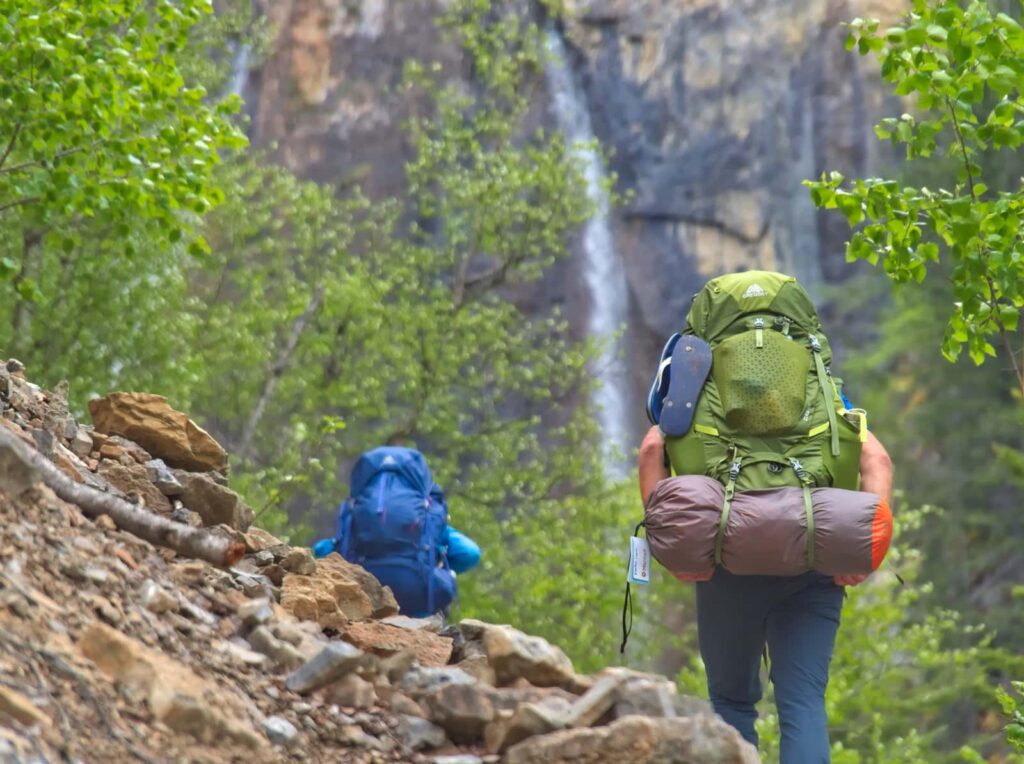 Backpackers hike up through trail with waterfall in the background.