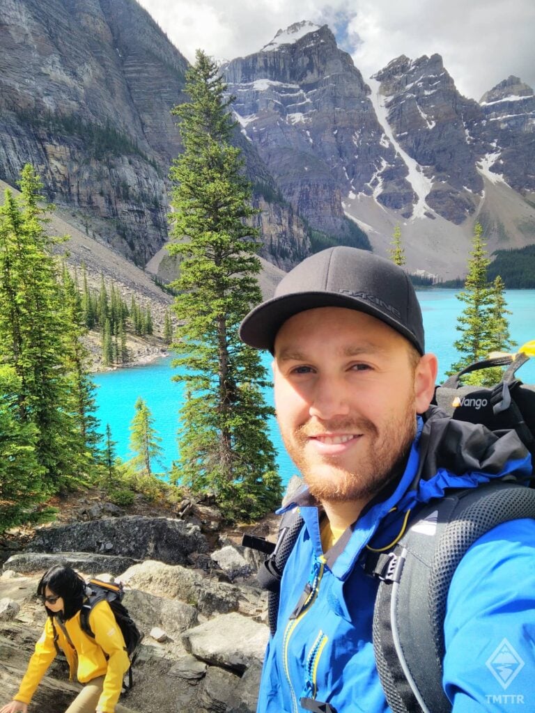 Man takes selfie in front of aquamarine lake and mountains.