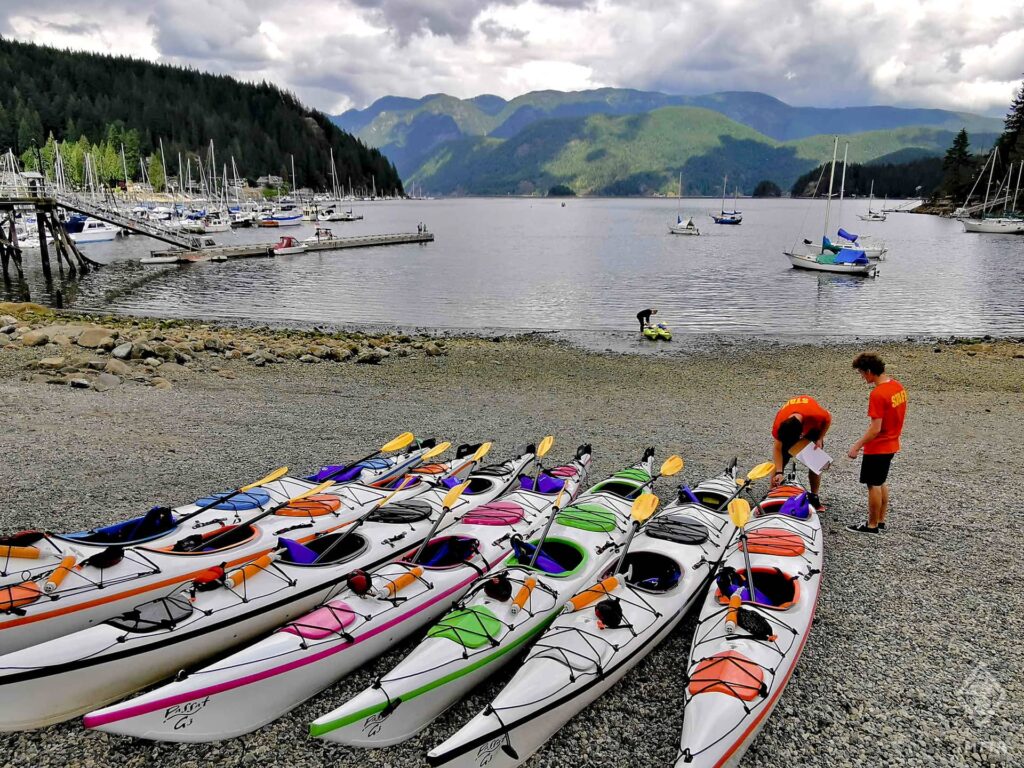 A selection of touring kayaks at Deep Cove, in front of the water.